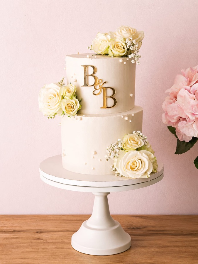 Tarta de dos pisos en marfil con rosas blancas, perlas e iniciales doradas