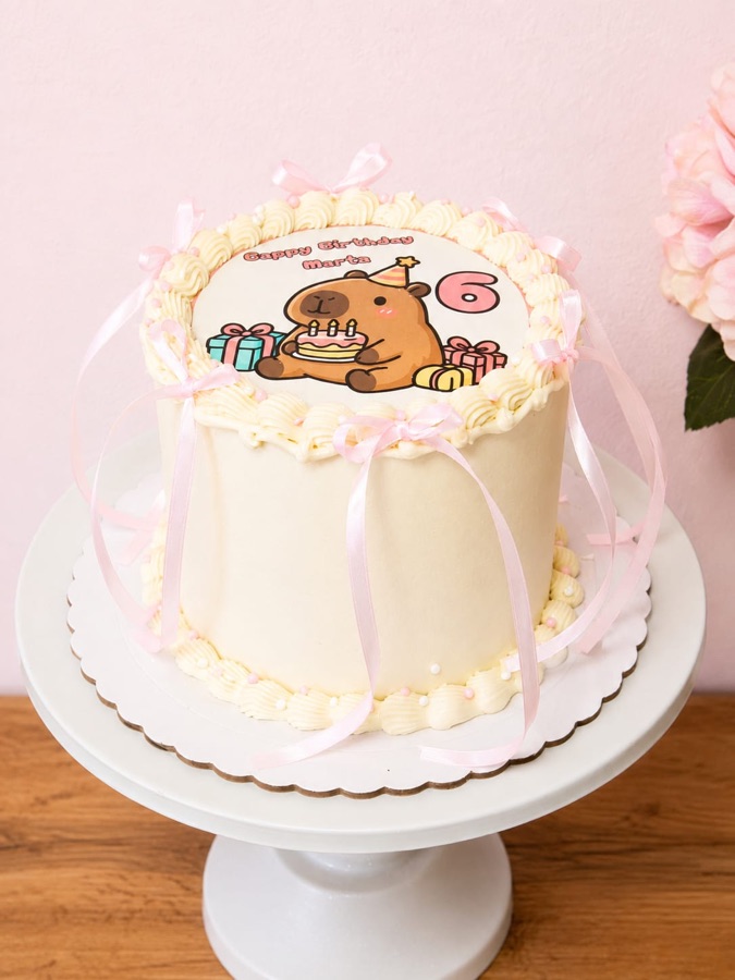 Tarta color crema con impresión de un capibara, lazos rosas y borde de nata