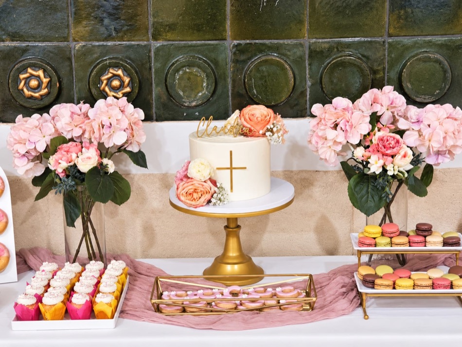 Mesa de comunión con tarta rematada por una cruz dorada y rosas, muffins con crema, macarons de colores y donuts glaseados